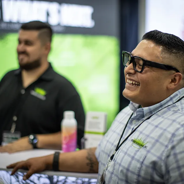 Smiling man in glasses and plaid shirt at a conference booth, with another staff member and promotional displays in the background.