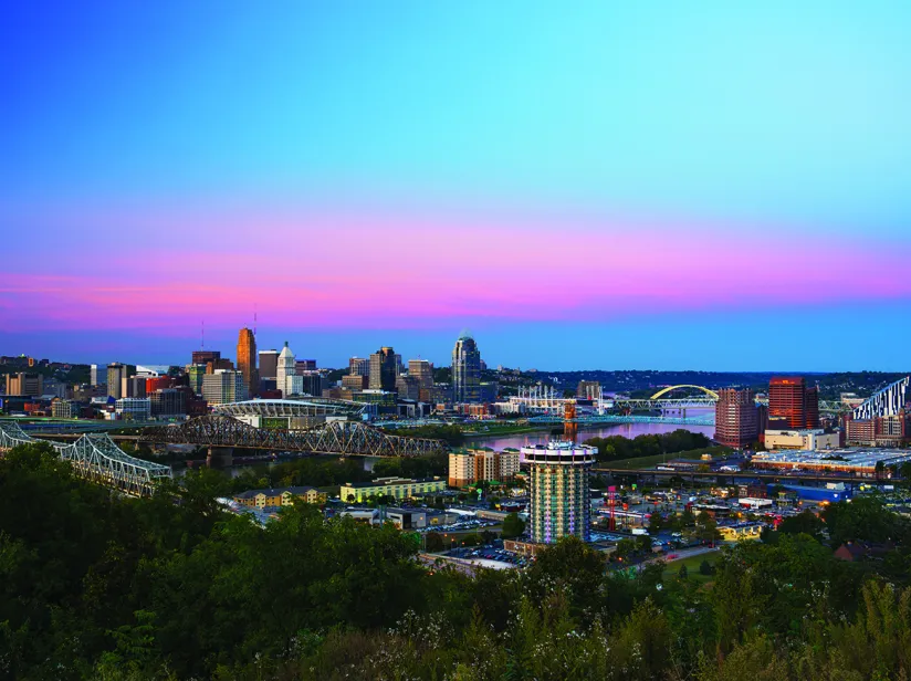 Cincinnati skyline at dusk with bridges over a river, colorful pink and blue sky, and surrounding urban and green areas.